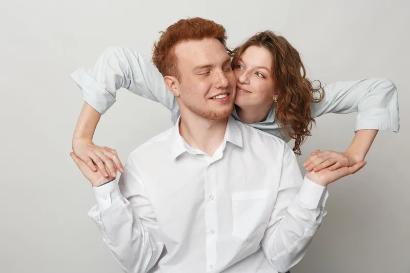 Young couple with red hair and curly hair wearing white shirts embraces and smiles in studio on grey background. Concept of love, relationship, happiness, and togetherness.