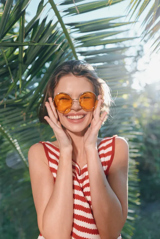 A cheerful woman wearing orange sunglasses and a red striped top smiles brightly among tropical foliage, creating a vibrant, sunny mood and a carefree summer vibe.