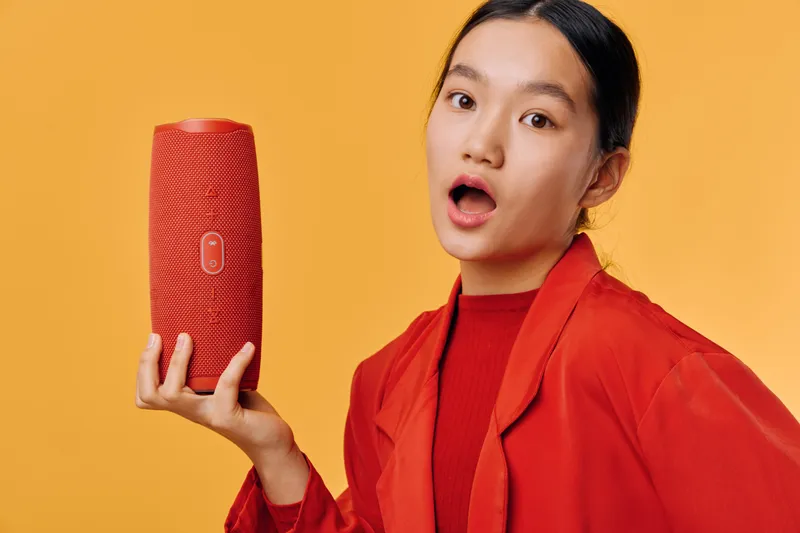 Woman holding wireless speaker with surprised expression on yellow background. Asian female in red jacket presenting portable audio device for music and sound technology.
