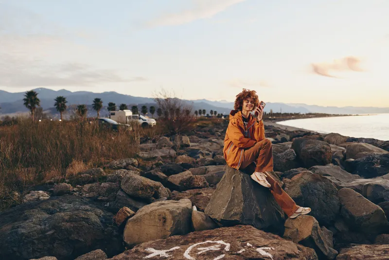 Woman lifestyle, seaside sunset, orange dress, sitting on rocks by the ocean, calm relaxed portrait, warm sunlight, coastal landscape with palms and distant mountains, evening sky clouds