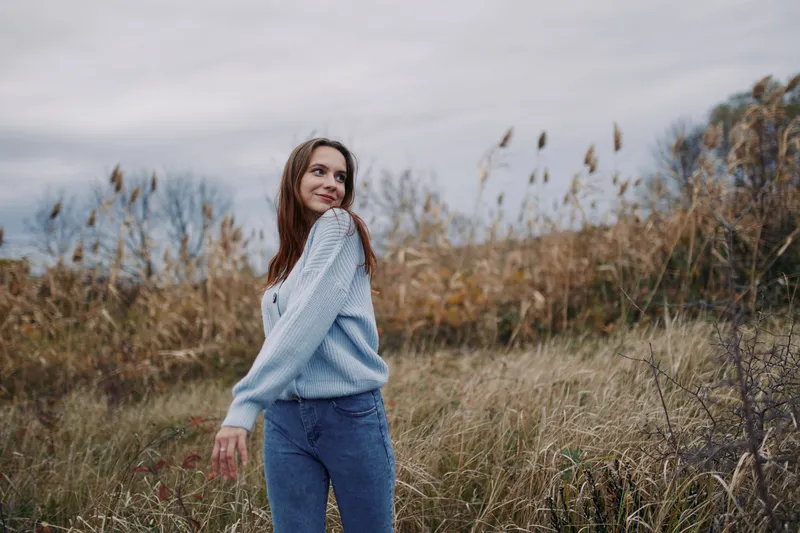 A relaxed portrait of a young woman in a cozy sweater walking through a grassy autumn field, radiating authenticity and natural confidence.