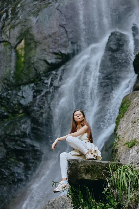 Young woman sitting on rock near large waterfall, nature background, casual outfit, serene mood, outdoor adventure, summer day.