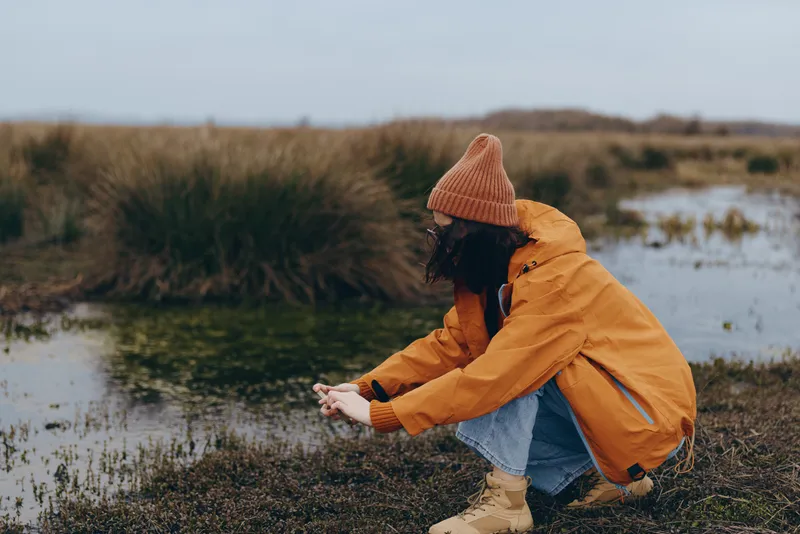 Woman lifestyle outdoors river autumn jacket hat walking nature concept, orange jacket and knit hat, person kneels by riverbank, smiling in calm scenic countryside, soft autumn light, candid travel