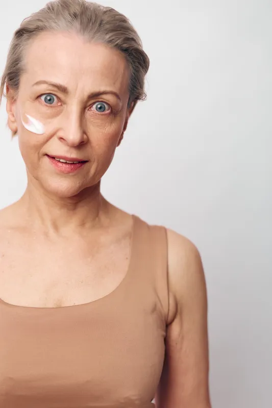 Mature woman with blue eyes applies white cream on cheek, wearing beige top. Close-up portrait on light background. Concept of skincare, aging, beauty routine, and self-care for older adults.
