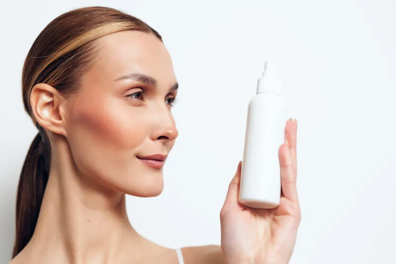 Focused portrait of a woman holding a white skincare bottle, clear skin, soft makeup, calm expression, studio lighting, minimal background, product emphasis, clean and fresh mood
