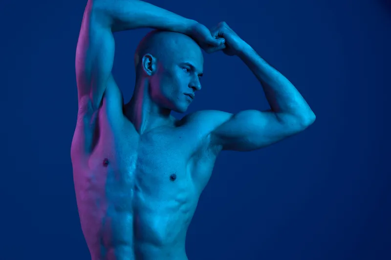 Athletic man posing with strong physique, bathed in blue light, showcasing muscles in a striking and captivating manner. Unique artistic expression in vibrant hues.