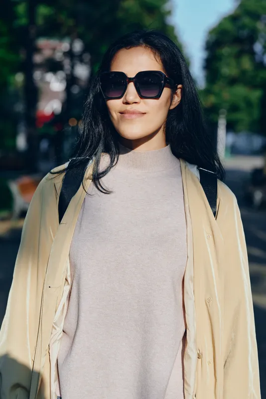 Woman wearing a cozy coat and oversized sunglasses walks along a sunlit street, showcasing casual fashion and urban style in a bright daytime moment