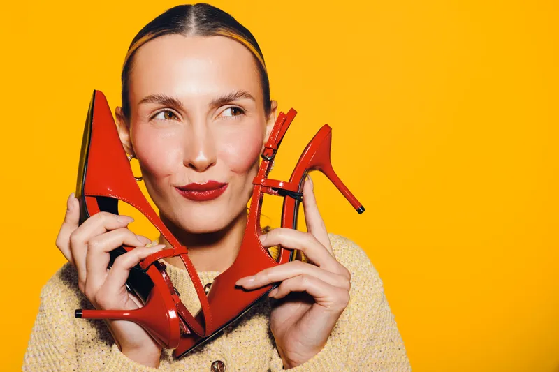 Young woman holds red high heels near face on bright yellow background. Fashion concept with playful expression and vibrant colors for style and footwear promotion.