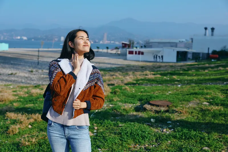 Woman in a knit jacket and scarf stands on a grassy coastal field, gazing upward with wind in hair, distant buildings and shoreline visible under a bright sky