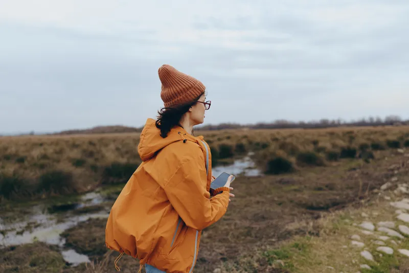 Woman outdoor lifestyle orange coat hat, standing in field, nature landscape, cloudy sky, slow walking posture, windy countryside path, calm mood