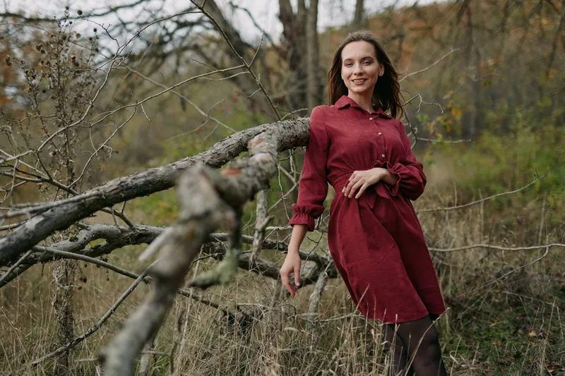 A woman in a deep red dress leans casually against a fallen tree in a wild meadow, natural pose and subtle smile conveying a sense of authenticity and credibility in a candid outdoor portrait.