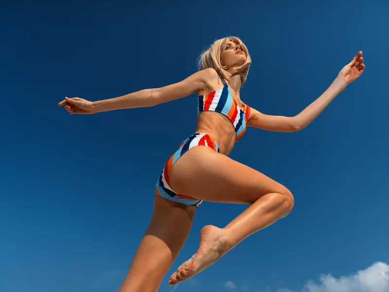 Athletic woman in a striped bikini leaps midair against a clear blue sky, capturing energy, balance, and carefree summer athleticism in a dynamic action shot.