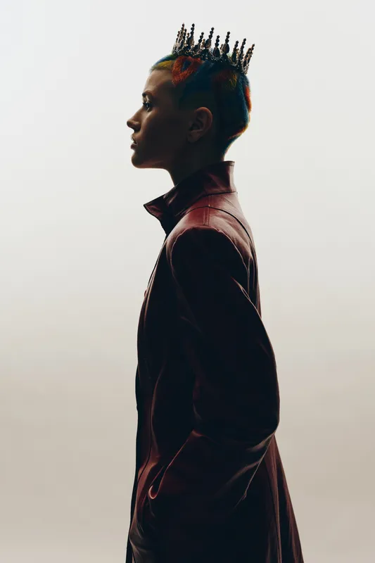 Profile of a young woman with a colorful braided hairstyle and a unique spiked crown accessory, wearing a dark leather jacket against a soft gradient background.