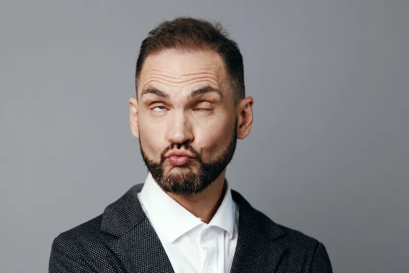 A professional man in a tailored suit strikes a playful pose with puckered lips, set against an isolated colored background for a business scene.