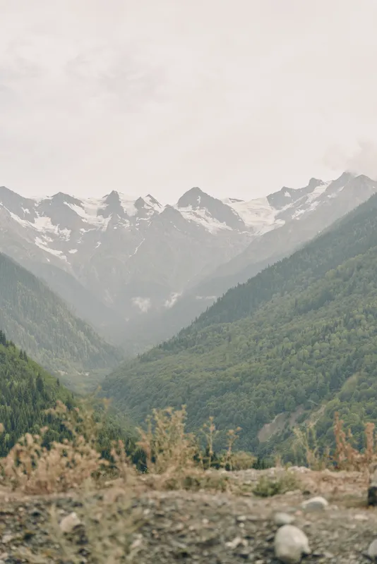 Majestic mountains rise over a deep valley with snow capped peaks, a rocky foreground and sparse vegetation creating a tranquil alpine landscape under soft light.