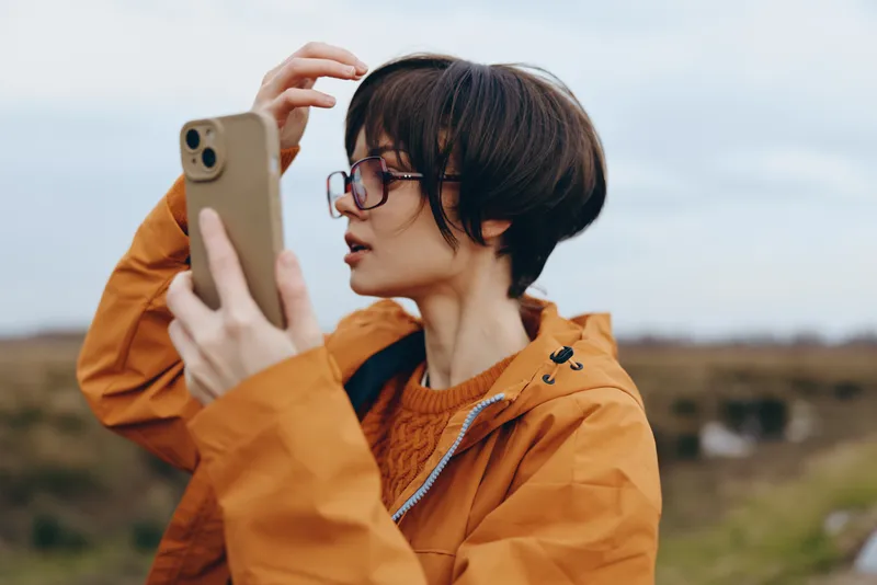 Woman lifestyle portrait outdoors in orange jacket, glasses on, holding a smartphone and taking a photo, daylight over rural fields, natural blurred background, relaxed confident mood