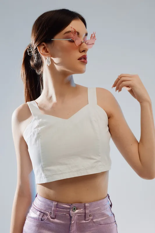 Young woman in a white crop top and purple pants poses with pink floral sunglasses. Her hair is tied back in a ponytail. Studio shot with soft lighting. Concept of beauty fashion.