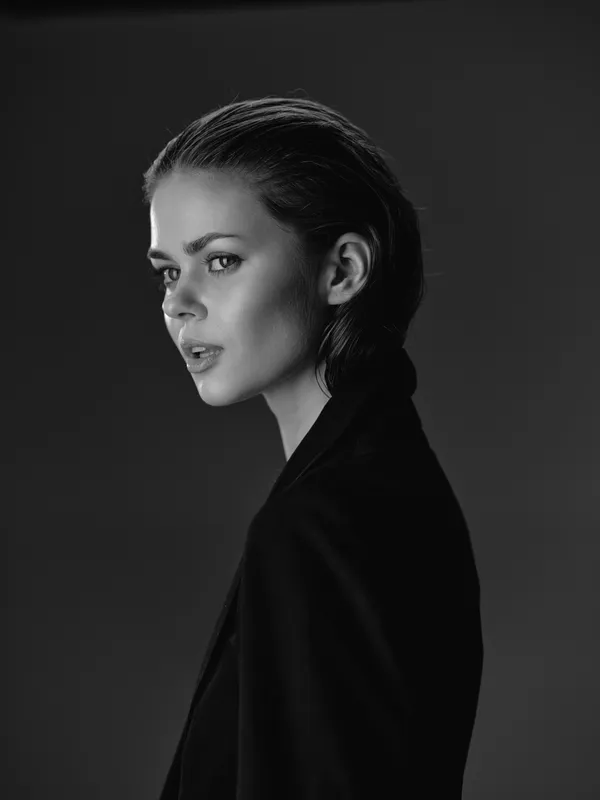 Black and white portrait of a serious woman with slicked-back hair wearing a dark jacket in studio lighting