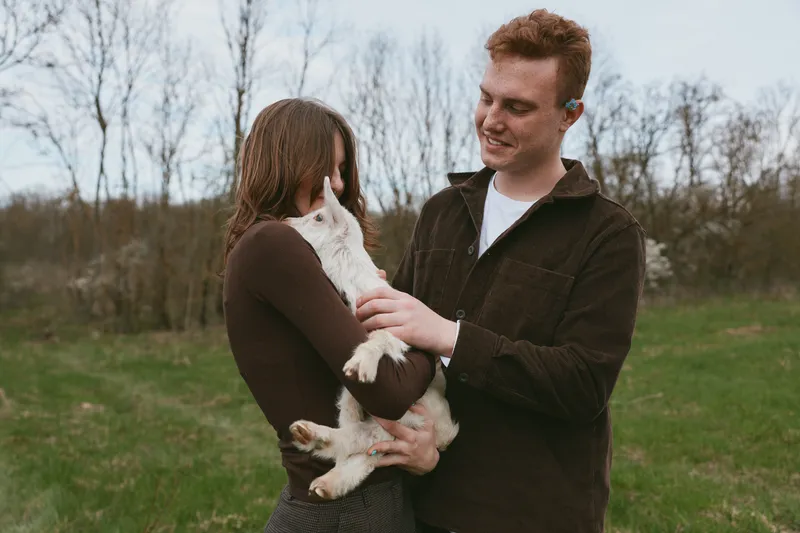 Happy couple outdoors with a goat kid, smiling and holding the animal in a grassy meadow, candid lifestyle moment showing warm affection and gentle connection