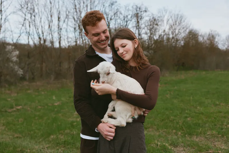 Young couple embraces tenderly while holding a small white goat outdoors in a green field during a calm overcast day, capturing a warm and natural lifestyle moment.