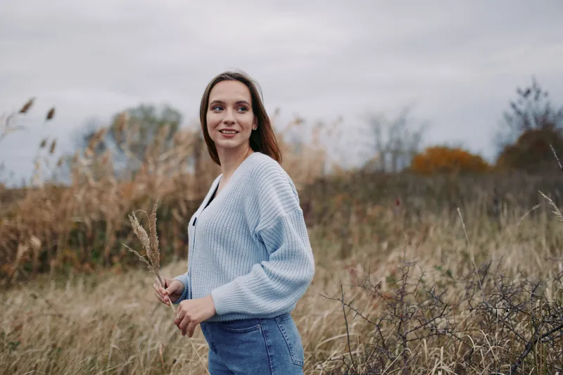 A candid portrait of a woman standing in a windswept field, conveying authenticity and quiet confidence. Natural light captures an honest expression and textured knitwear detail.