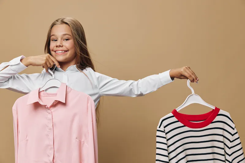 Girl choosing clothes holding pink shirt and striped sweater on hangers with smile. Fashion decision, wardrobe selection, casual outfit, youth and style concept.