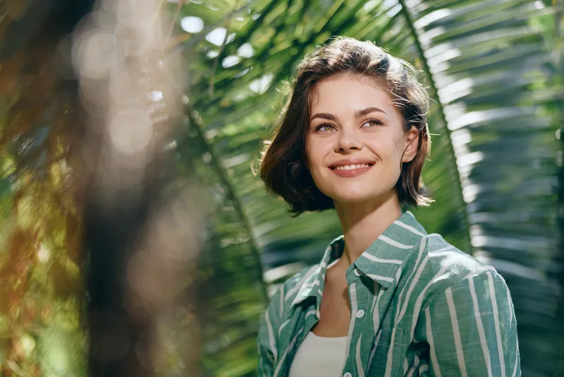 Woman, smile, outdoor portrait in natural light. A joyful model stands amid lush greenery, wearing a green striped shirt, exuding warmth, confidence and relaxed charm in sunny surroundings, natural