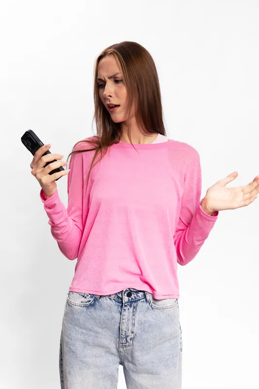 Young woman in pink sweater looks confused at a smartphone, holding it in one hand and gesturing with the other. Concept of frustration, communication, and technology issues.
