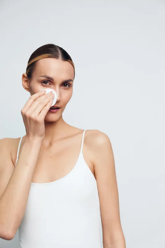 Young woman with a cotton pad wiping her face in a clean studio, practicing skincare with soft lighting and a calm, minimal mood.