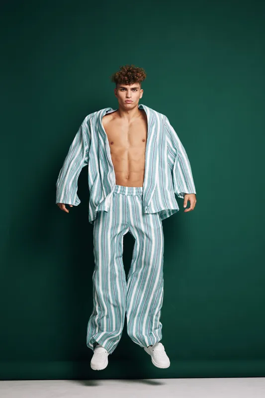 Young man jumping in striped loose pants and open shirt with bare chest against green background. Casual fashion, confident pose, studio portrait with modern style and athletic body.