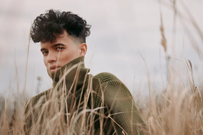 Moody outdoor portrait of a young man in a chunky green sweater among tall dry grass, capturing a candid natural expression that emphasizes authenticity and visual credibility in a realistic scene.