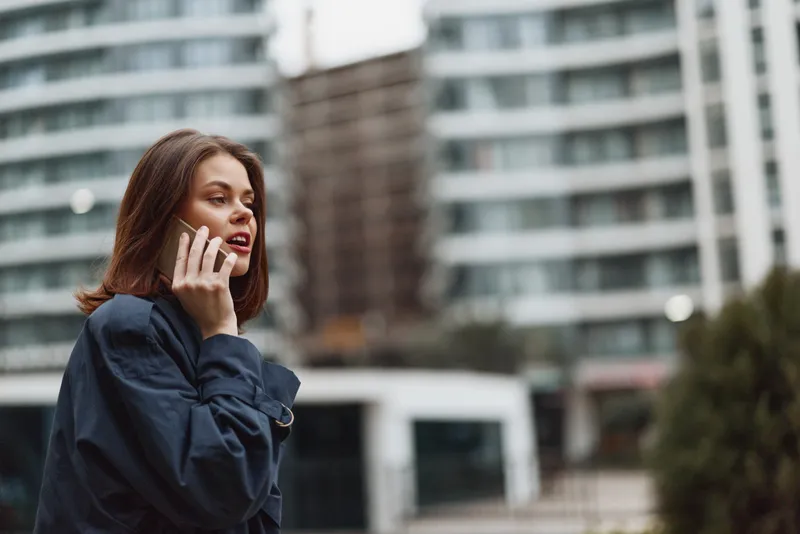 Young woman in a denim jacket talks on her smartphone outdoors in an urban setting, focused during a daytime call with a blurred city backdrop and cool tones.