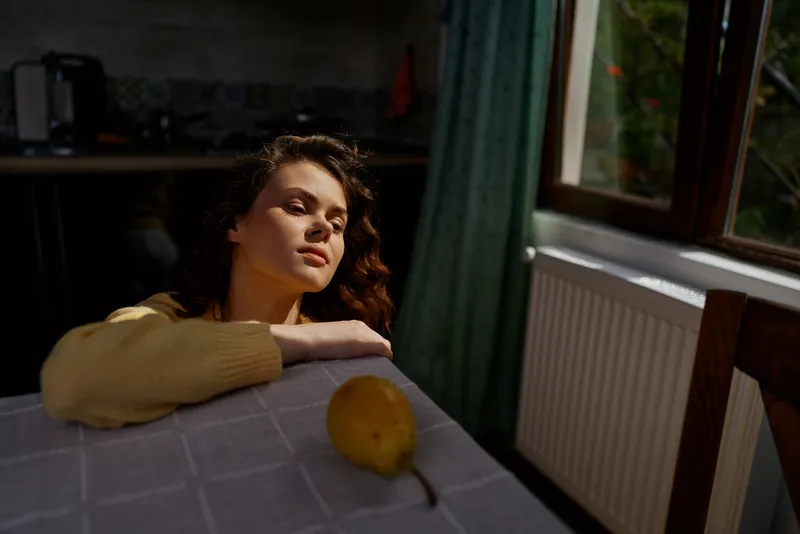 woman at home, contemplative and still, bathed in sunlight during the afternoon, seated at a table by a window with an orange nearby, evoking calm, reflective mood.