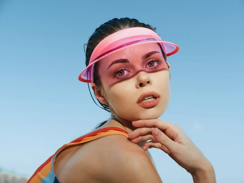 Portrait of a young woman wearing a pink visor and pink tinted eye protection, posing against a bright blue sky with warm sunlight and a relaxed, confident expression.