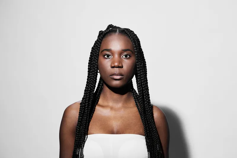 Portrait of a young Black woman with braided hair, wearing a strapless top, expressing confidence against a minimalist grey background, ideal for beauty and fashion concepts