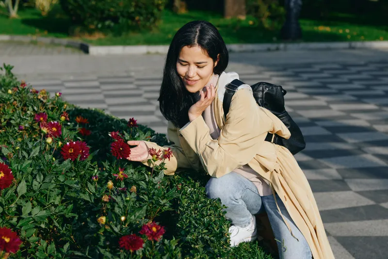 Woman kneeling beside vibrant flowers, examining petals in a sunny park, wearing light jacket and backpack, a candid outdoor moment of exploration and delight