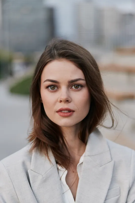 Confident businesswoman posing outdoors in a modern urban environment, showcasing professionalism and poise, wearing a light gray blazer and white blouse, with a soft expression.