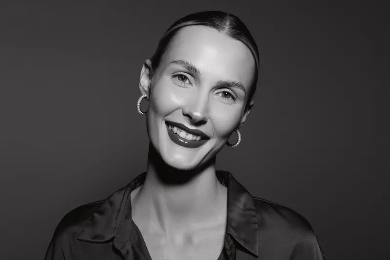 Portrait of a smiling white woman with fair skin and slicked-back hair wearing hoop earrings and a dark shirt, isolated on a dark background. Concept of happiness and elegance.