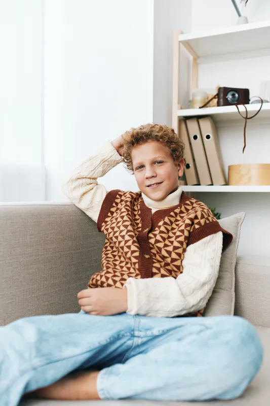 Young boy sits relaxed on a sofa indoors, wearing a patterned knit vest and cozy sweater, smiling softly as he rests his arm on the back of the couch.