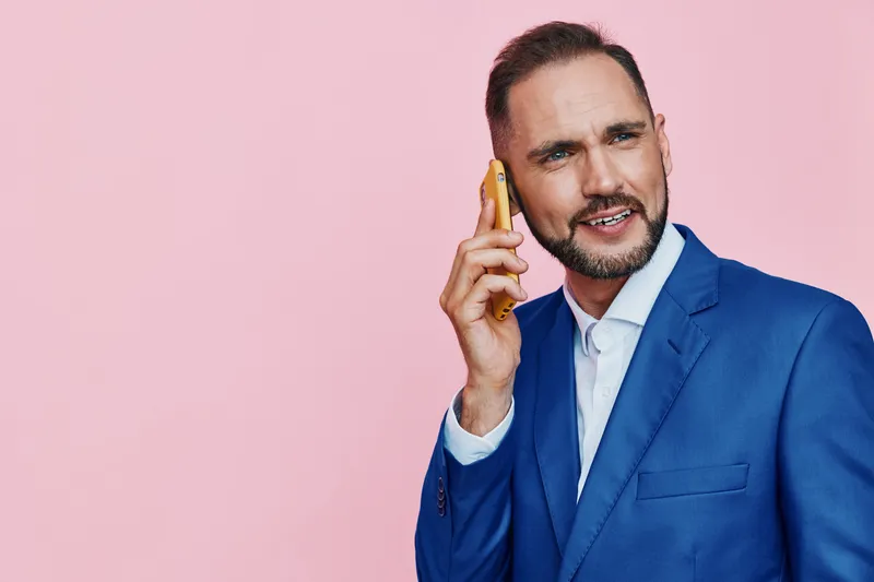 A professional man in a blue suit speaks on a phone against a solid pink background, projecting confidence, focus, and strong emotions in a corporate setting.