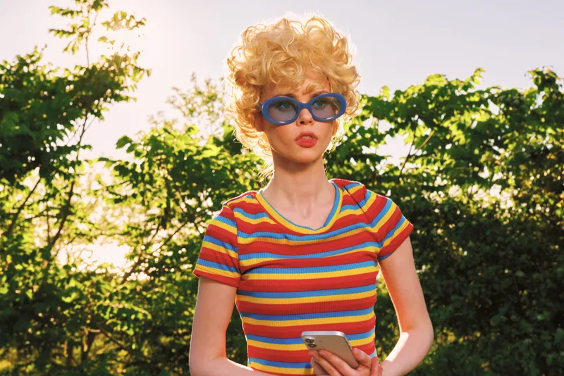 Smiling woman with curly hair wearing a striped colorful shirt and blue sunglasses outdoors. Casual lifestyle, retro vibes, bright light and cheerful mood in a sunny park setting.