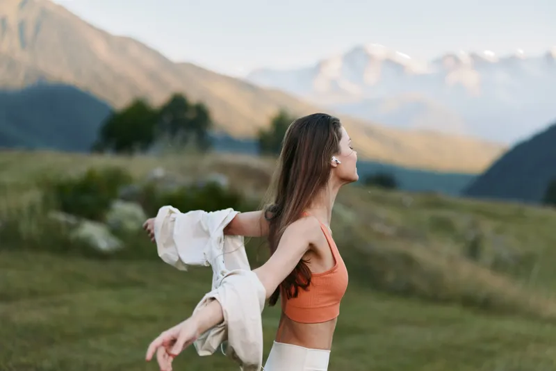 A cheerful outdoor-inspired portrait captures a young woman in a scenic mountainous landscape, arms open, sunlight on her skin, radiating freedom and fresh adventure.