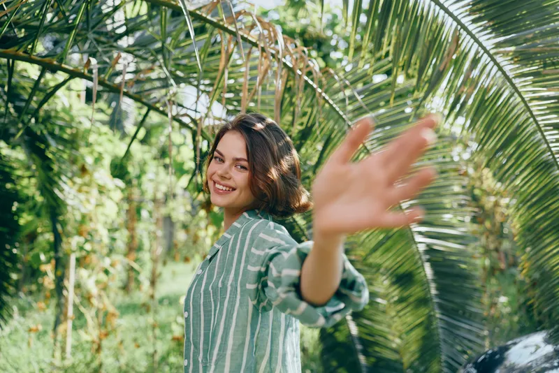 Cheerful woman in a striped shirt extends a friendly wave in a lush garden setting, sunlight filtering through tropical palm leaves.