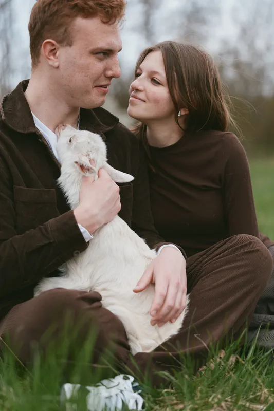 Young couple sitting on grass with a white baby goat in their arms enjoying a calm moment outdoors in casual clothes during a relaxed lifestyle scene.
