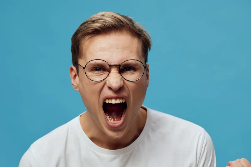 Angry woman with short brown hair and glasses screaming against a plain blue background, expressing frustration or anger, studio portrait, emotional human face.