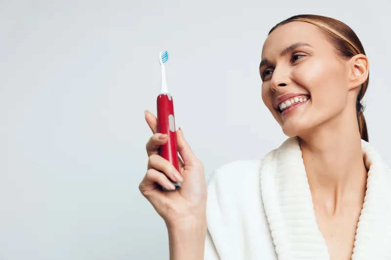 Smiling woman holding electric toothbrush with blue bristles. Close-up on light background. Concept of dental care, hygiene, and healthy lifestyle. Soft natural lighting highlights joyful mood.