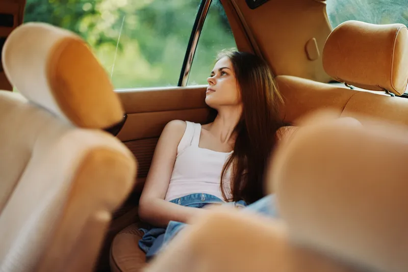 Young woman with long hair in a car, looking thoughtfully out the window, wearing a casual white tank top and denim shorts, capturing a moment of contemplation in nature.