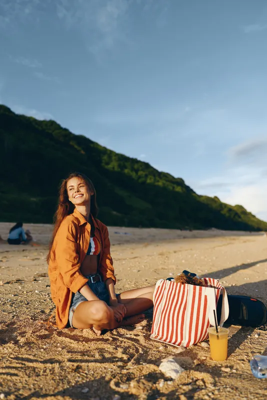Joyful woman sits on a sandy beach under warm sun, wearing an orange shirt, with striped tote and picnic items nearby, coastal cliffs in the background, relaxed vacation mood
