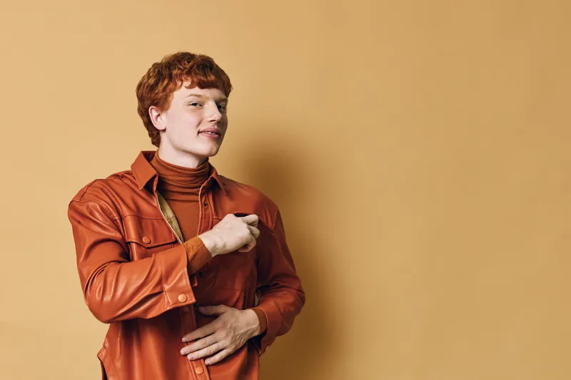 Young man in orange shirt and brown turtleneck posing with confident expression on beige background. Red hair, casual style, studio portrait with warm tones and relaxed mood.