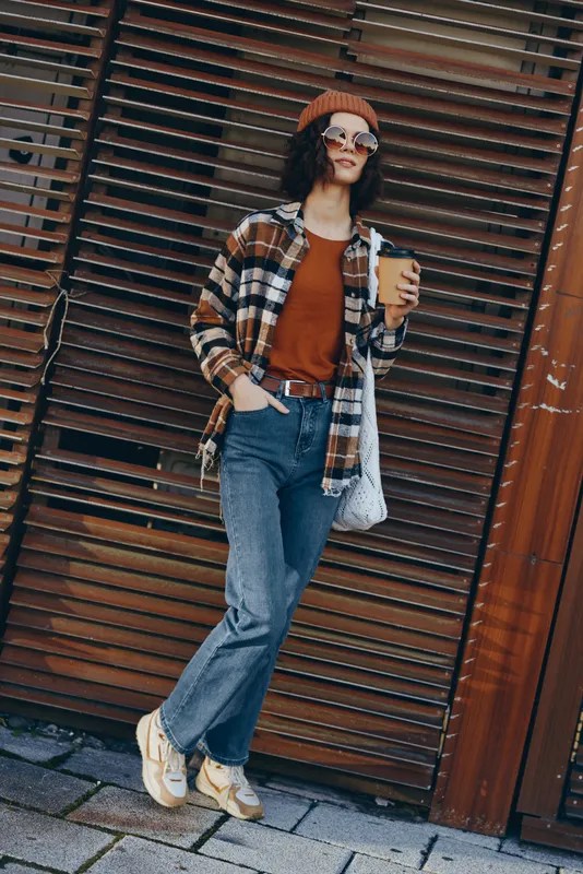 Woman holding coffee in urban street, casual outfit with sunglasses, beanie, flannel and jeans leaning against shutter; candid lifestyle portrait with authenticity and golden hour glow.